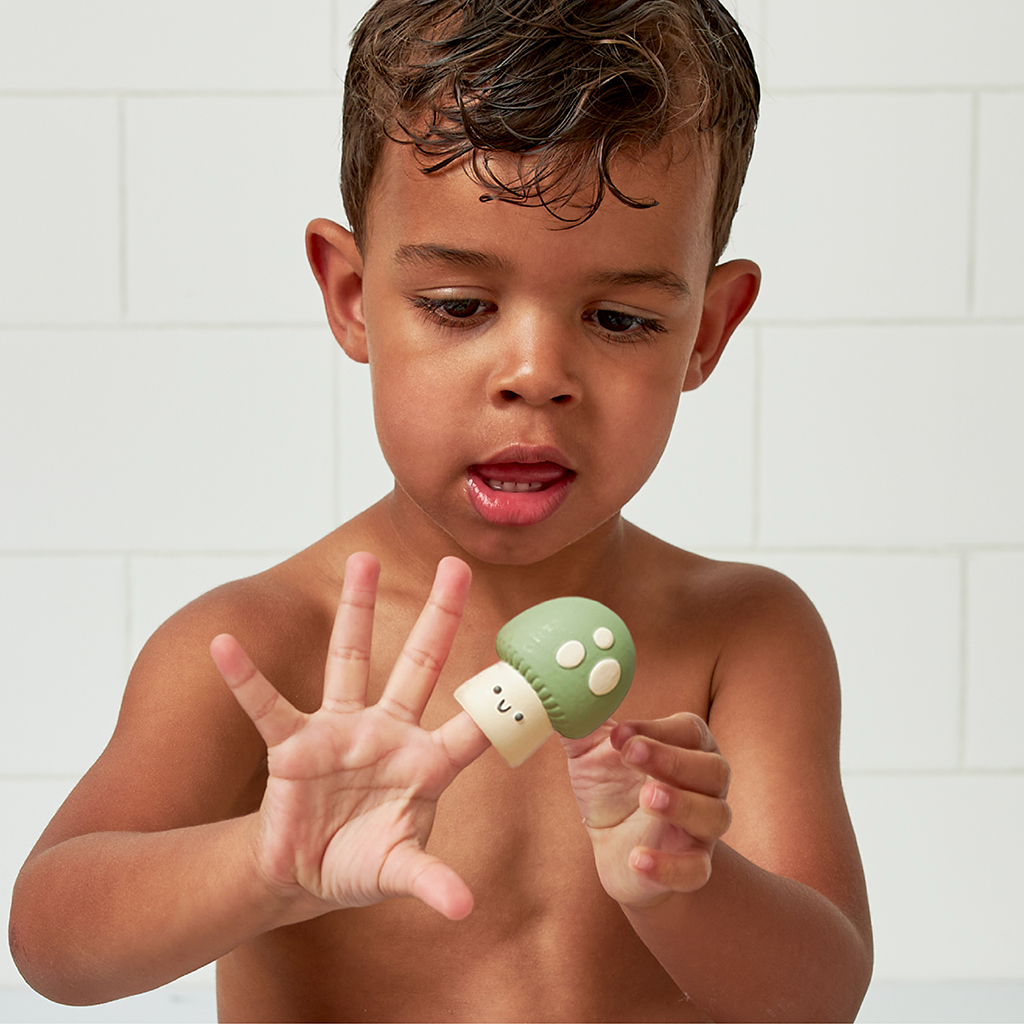 FP8560_Itzy_Ritzy_Bath_Itzy_Bitzy_Finger_Puppets A young child with wet hair holds up and examines a green Itzy Ritzy Finger Puppet Bath Toy shaped like a mushroom in front of a white tiled background.