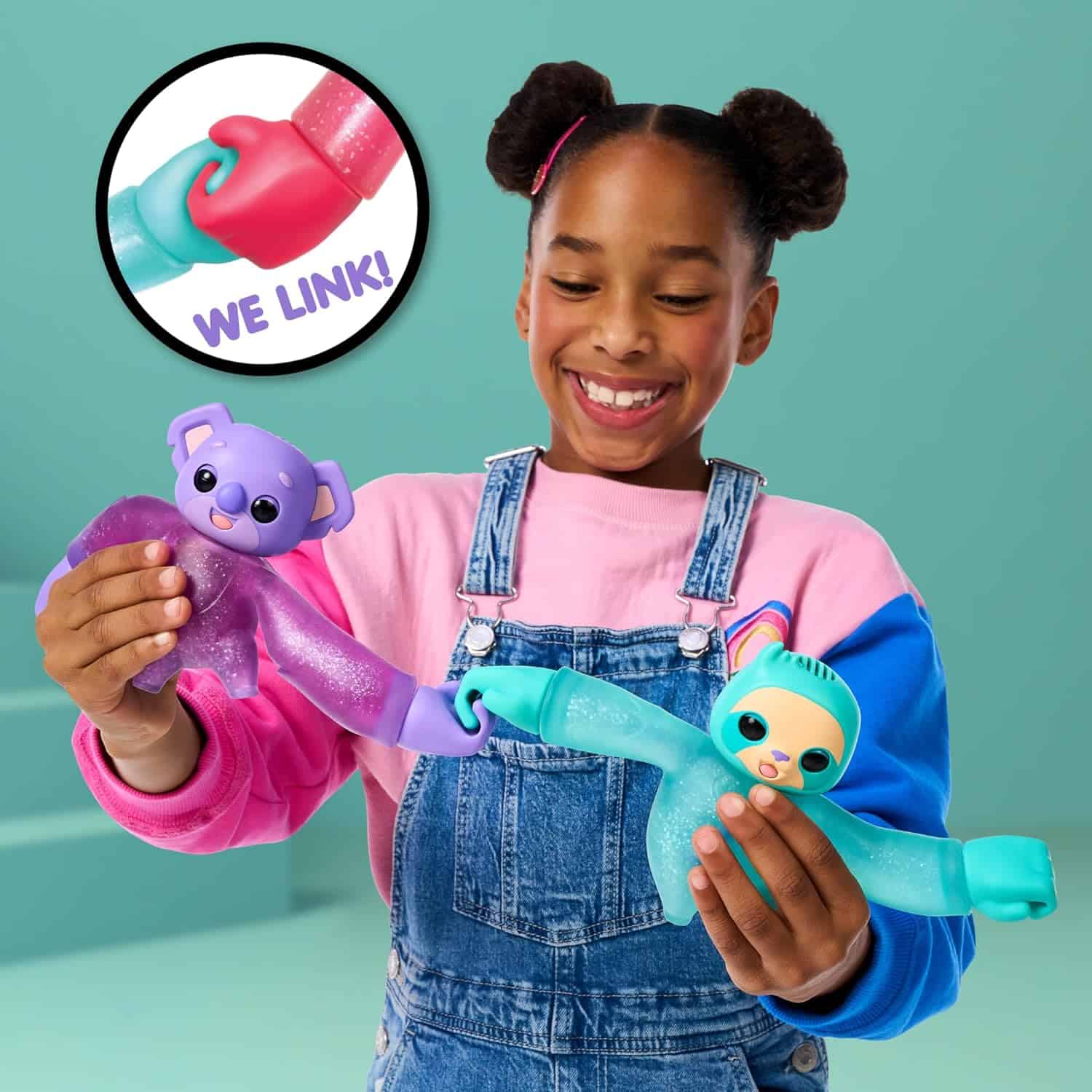 Little Live Pets Hug n' Hang Zoogooz - Koomi Koala. an Interactive Electronic Squishy Stretchy Toy Pet with 70+ Sounds & Reactions. Stretch, Squish and Link Their Hands (7) A child in a pink shirt and denim overalls is playing with two colorful toy animals that link hands. A close-up inset shows how the toys connect, with text that reads "WE LINK!.