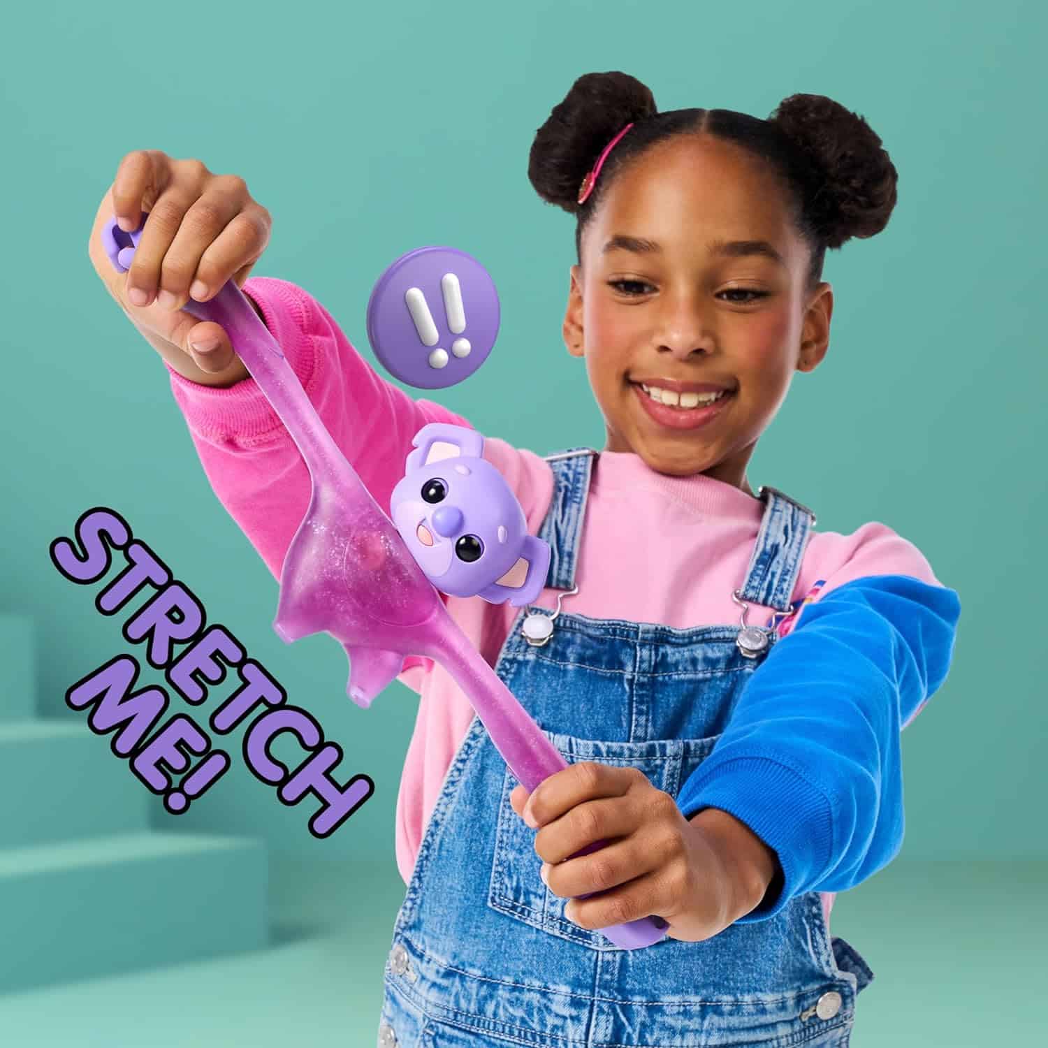 Little Live Pets Hug n' Hang Zoogooz - Koomi Koala. an Interactive Electronic Squishy Stretchy Toy Pet with 70+ Sounds & Reactions. Stretch, Squish and Link Their Hands (8) A child with two buns in her hair stretches a long, purple toy with a cute animal face and ears. The text "STRETCH ME!" is displayed on the left side. The background is light green.