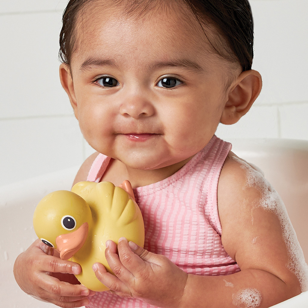 RD8553_Itzy_Ritzy_Bath_Itzy_Ducky_Family A baby wearing a pink tank top holds an Itzy Ritzy Ducky Family Floating Bath Toy while sitting in a bathtub.