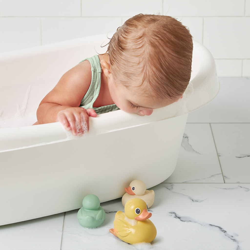 RD8553_Itzy_Ritzy_Bath_Itzy_Ducky_Family A baby in a bathtub leans over the side to look at three Itzy Ritzy Ducky Family Floating Bath Toys on the tiled floor next to the tub.