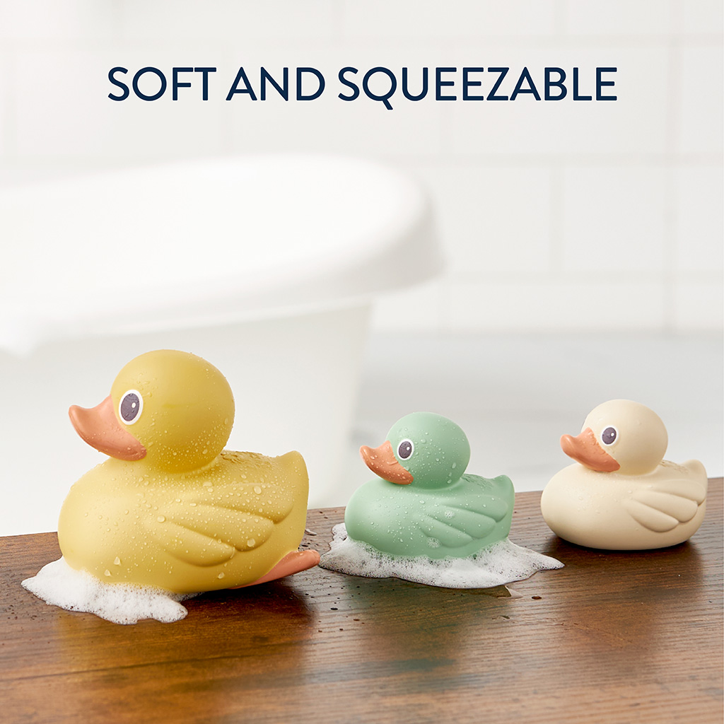 RD8553_Itzy_Ritzy_Bath_Itzy_Ducky_Family Three Itzy Ritzy Ducky Family Floating Bath Toys in yellow, green, and white sit on a wooden surface with soapy bubbles. A bathtub is visible in the background. Text above reads "Soft and Squeezable.