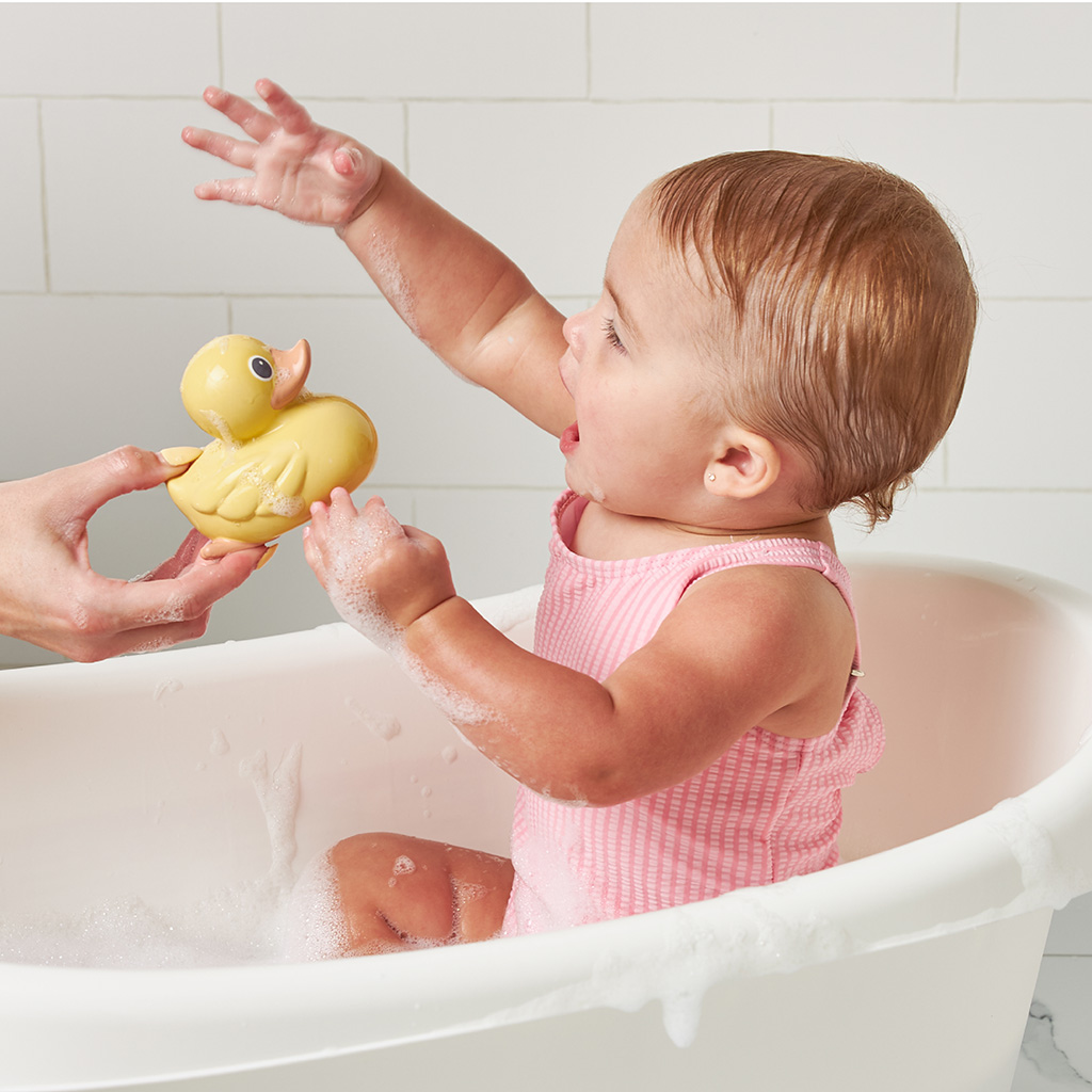 RD8553_Itzy_Ritzy_Bath_Itzy_Ducky_Family A baby in a pink bathing suit sits in a bathtub, reaching excitedly for an Itzy Ritzy Ducky Family Floating Bath Toy held by an adult's hand.
