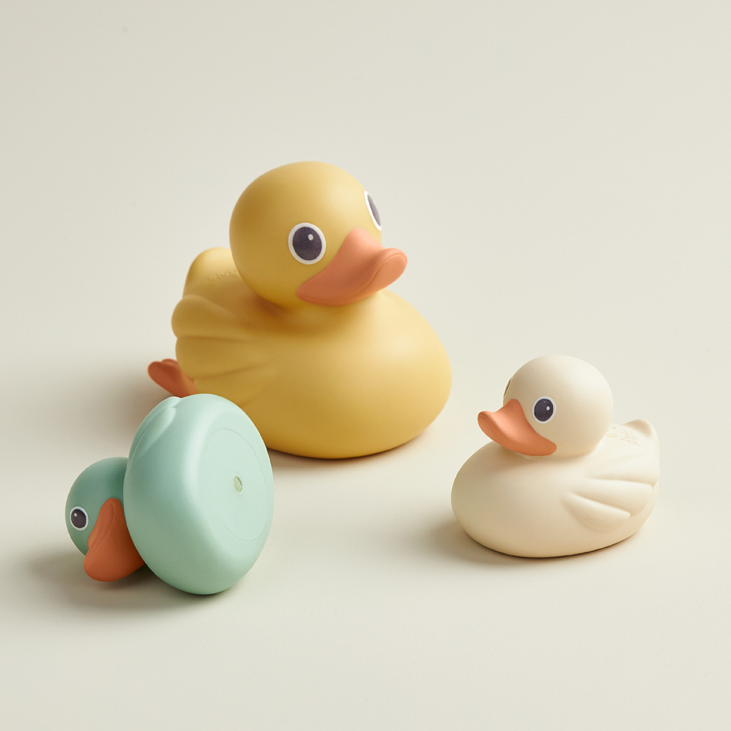 RD8553_Itzy_Ritzy_Bath_Itzy_Ducky_Family Three toys from the Itzy Ritzy Ducky Family Floating Bath Toys set are placed on a light-colored surface: one yellow, one white, and one green with its head turned.