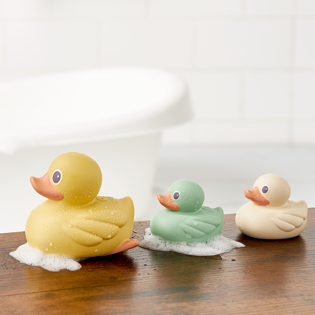 RD8553_Itzy_Ritzy_Bath_Itzy_Ducky_Family Three Itzy Ritzy Ducky Family Floating Bath Toys—yellow, green, and white—are lined up on a wooden surface with soap bubbles, with a bathtub in the blurred background.