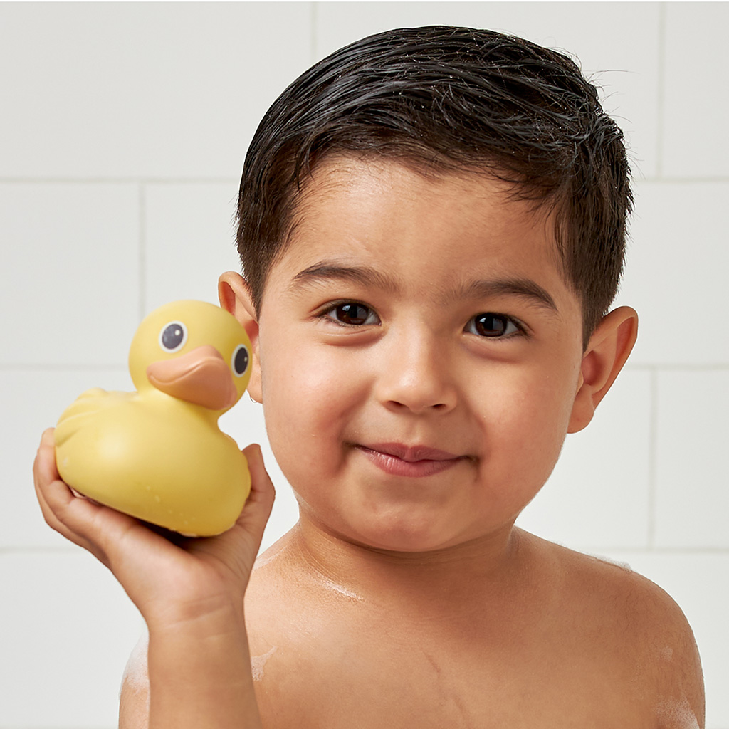 RD8553_Itzy_Ritzy_Bath_Itzy_Ducky_Family A young child with wet hair holds an Itzy Ritzy Ducky Family Floating Bath Toy, standing in front of a white tiled background.