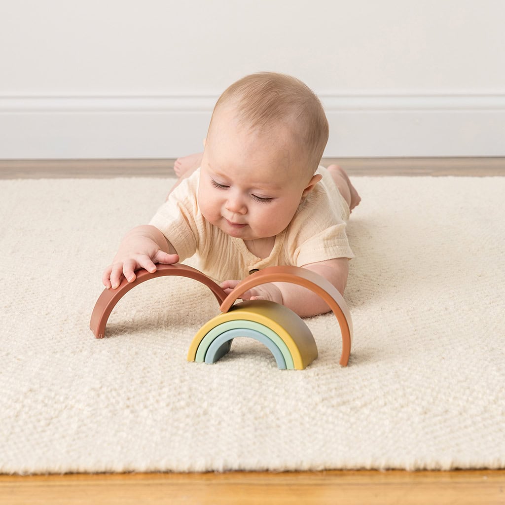 RS8548 Itzy Ritzy Rainbow Stacking Toy (1) Baby lying on a rug playing with a wooden rainbow toy.
