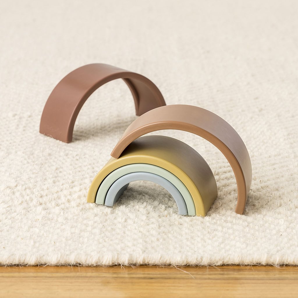RS8548 Itzy Ritzy Rainbow Stacking Toy (8) Wooden rainbow toy pieces in pastel colors, stacked in an arch shape on a cream carpet.
