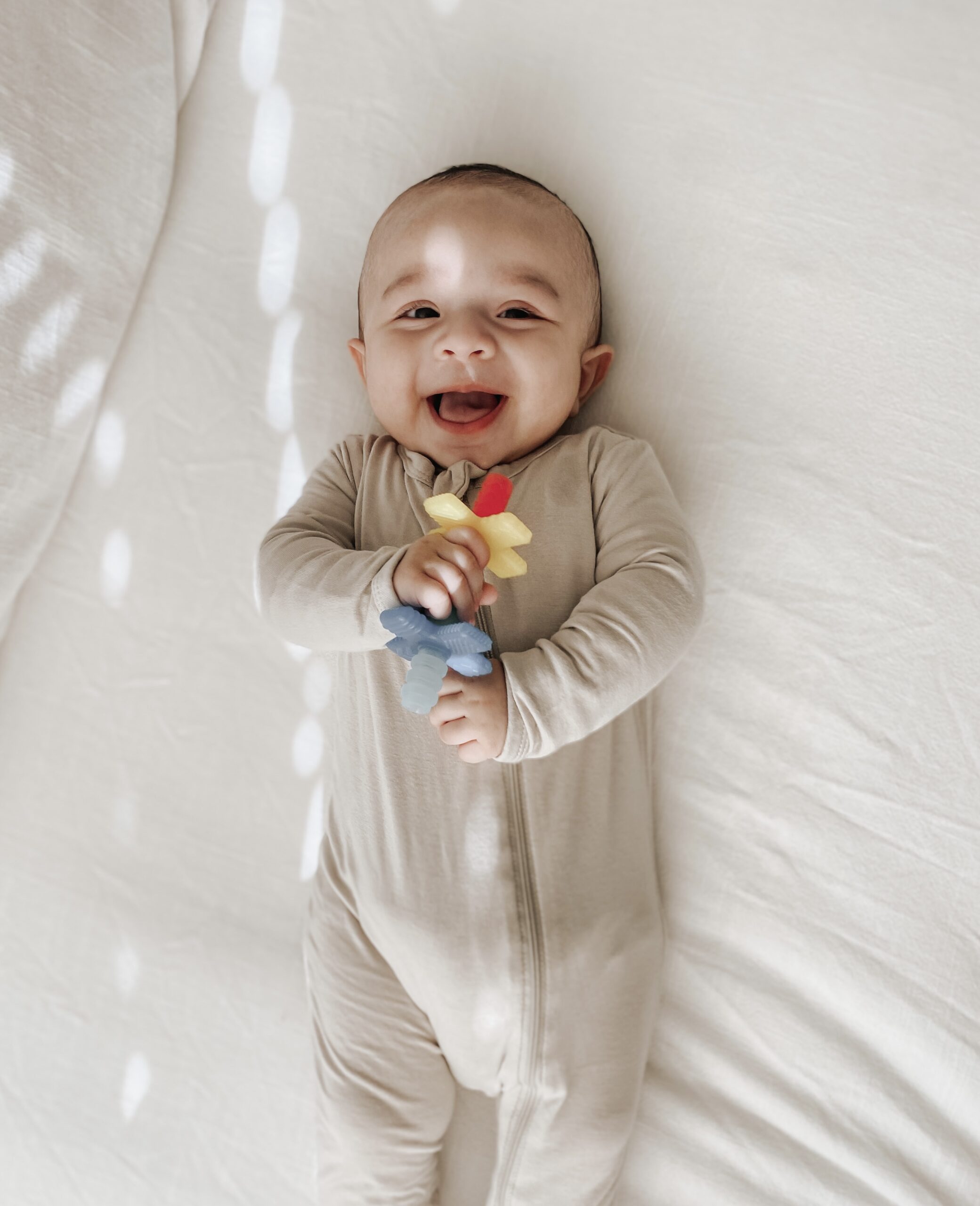 A baby in a beige onesie lays on a bed while smiling and holding a colorful toy flower.