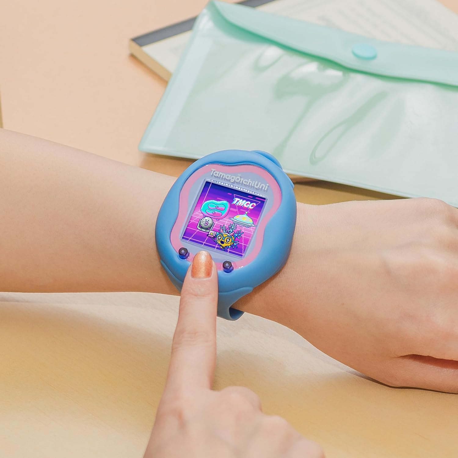 Tamagotchi Uni - Blue (9) A person interacts with a blue Tamagotchi smartwatch displaying a colorful screen, while resting their hand on a light-colored surface. A translucent green envelope is partially visible in the background.