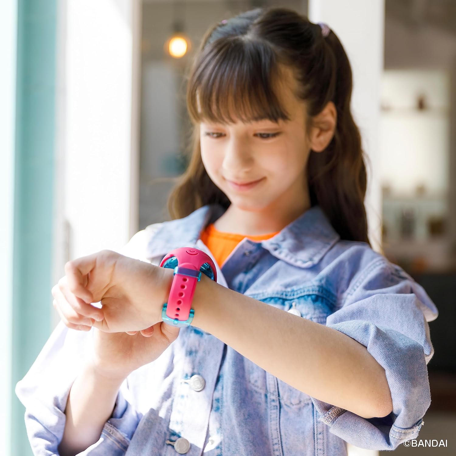 Tamagotchi Uni - Pink (2) A young girl in a denim jacket looks at her wrist wearing a Tamagotchi Uni smartwatch in a brightly lit room.