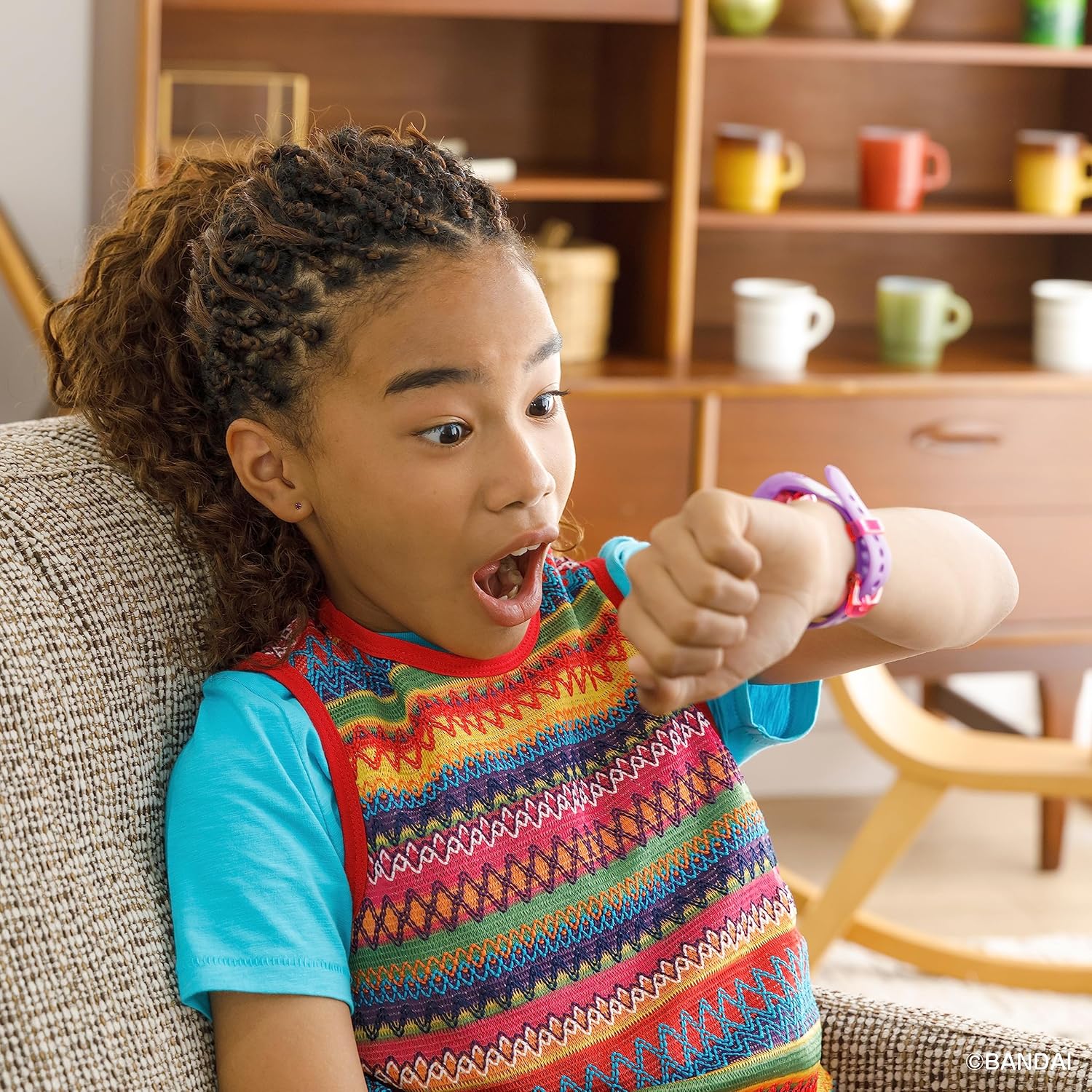 Tamagotchi Uni - Purple (4) A young child with braided hair looks surprised while glancing at their wrist, which has a colorful watch. The child is indoors, sitting in a living room with shelves and colorful mugs in the background.