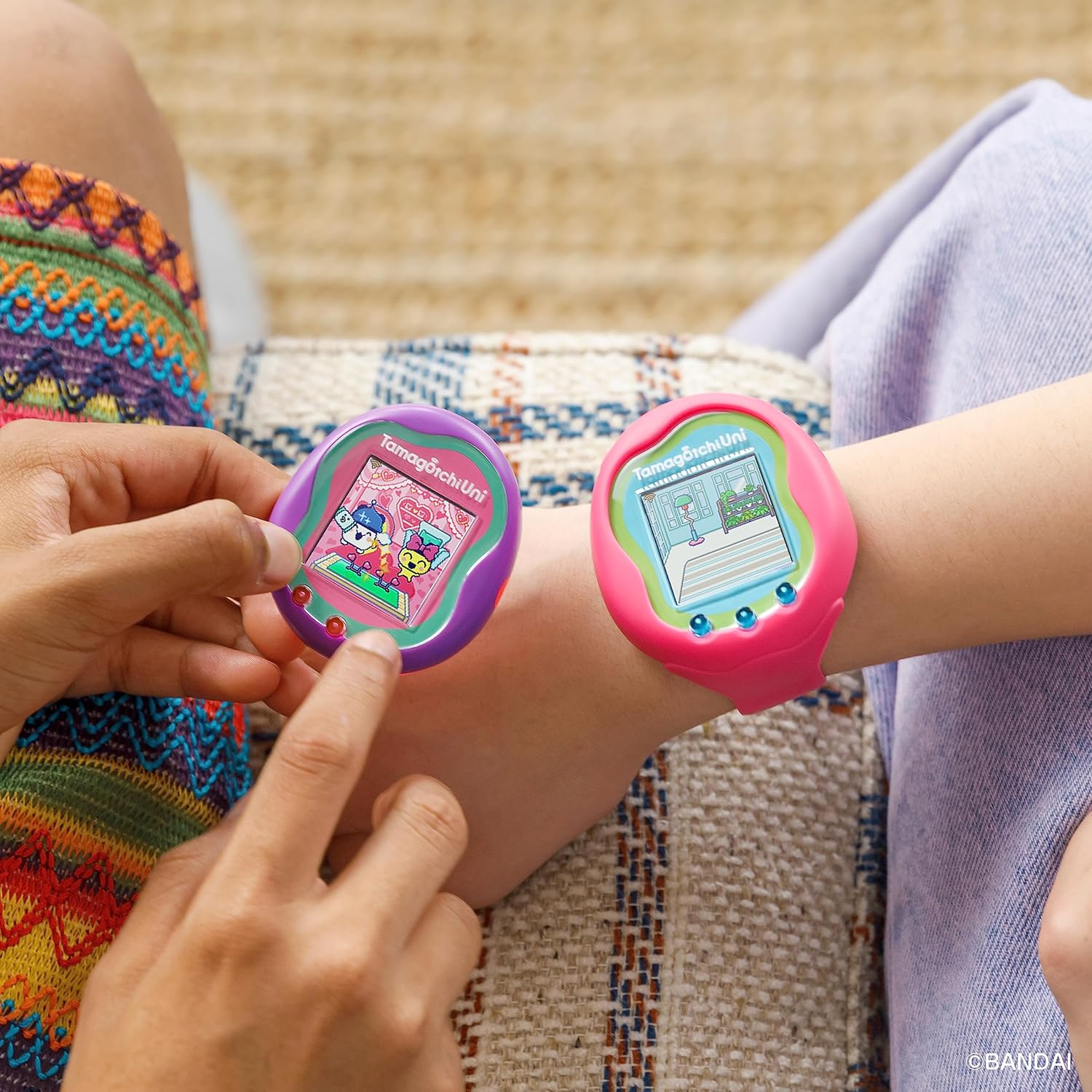 Tamagotchi Uni - Purple (8) Two people interacting with pink Tamagotchi Uni wearable devices on their wrists, one person using finger to touch the screen. Both devices display digital pets.