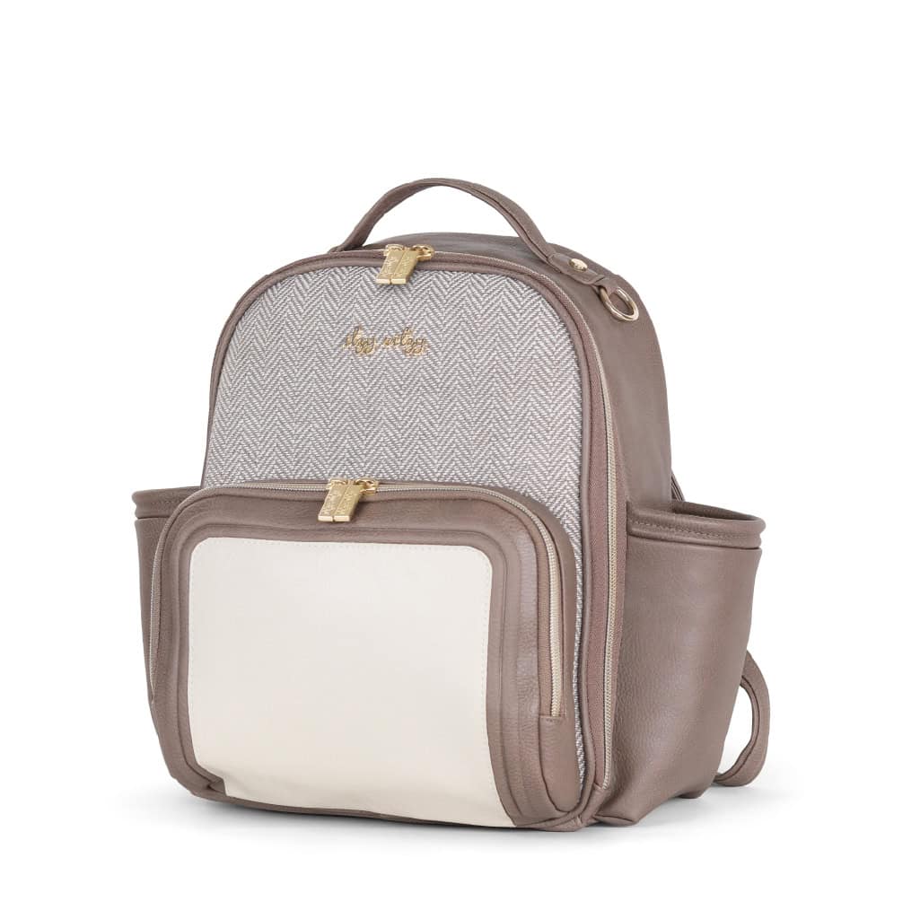 itzy-ritzy-101401590-itzy-mini-plus-backpack-vanilla-latte-1 A beige and white backpack with gold zippers and a small front pocket. The bag has a handle on top and two side pockets.