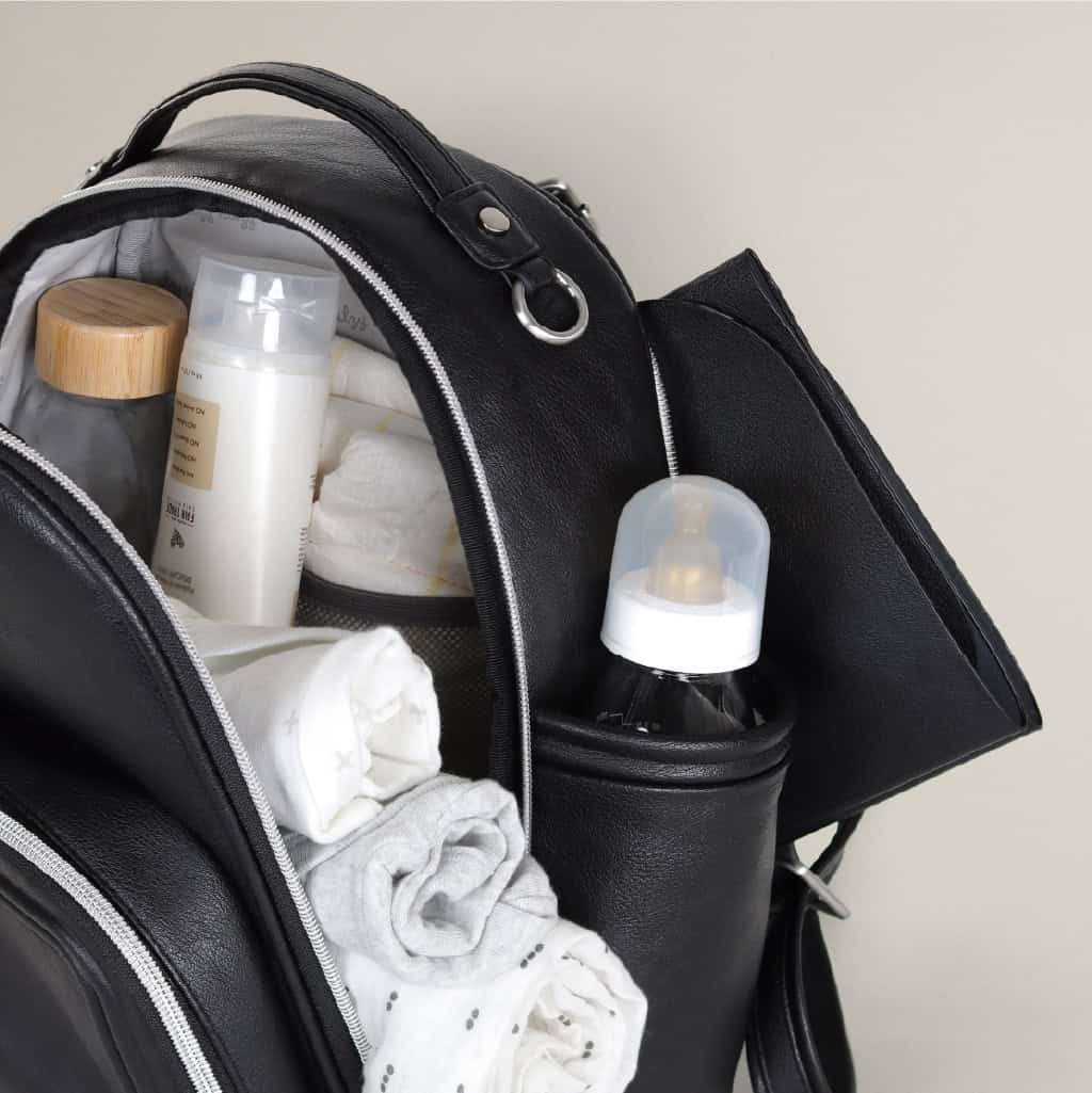 itzy-ritzy-101401619-itzy-mini-plus-backpack-noir-6 An open black backpack showing a baby bottle, rolled-up clothes, a wooden container, and a bottle with white liquid inside.