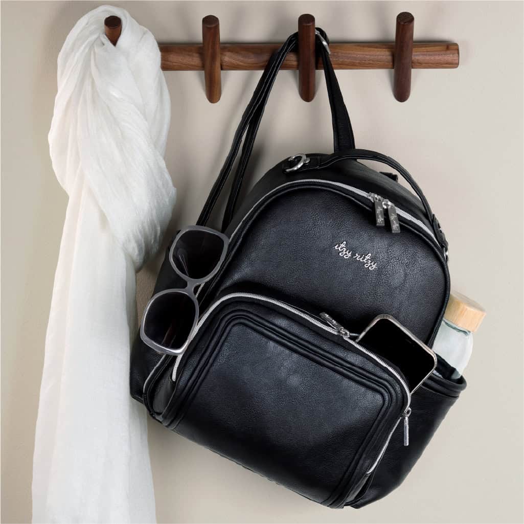 itzy-ritzy-101401619-itzy-mini-plus-backpack-noir-9 A black leather handbag hangs on a wooden coat rack beside a white scarf. The bag has a front zipper pocket, and contains a pair of sunglasses, a smartphone, and a water bottle in its side pocket.