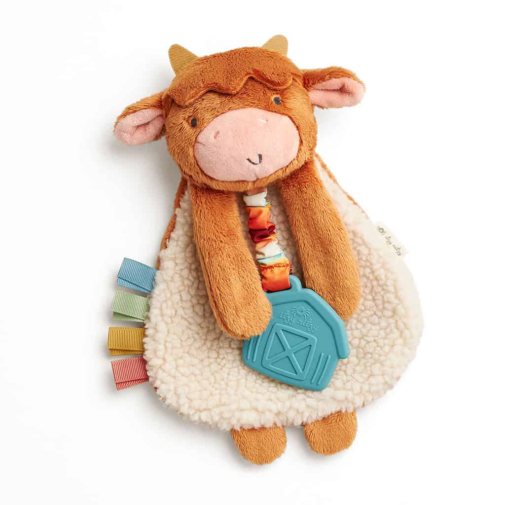 302801668_Itzy_Ritzy_Itzy_Lovey_Highland_Cow A plush toy shaped like a cow, with a soft, furry body and colorful taggy ribbons on its side. The toy features a pacifier clip with a blue, house-shaped teether in its front pocket.