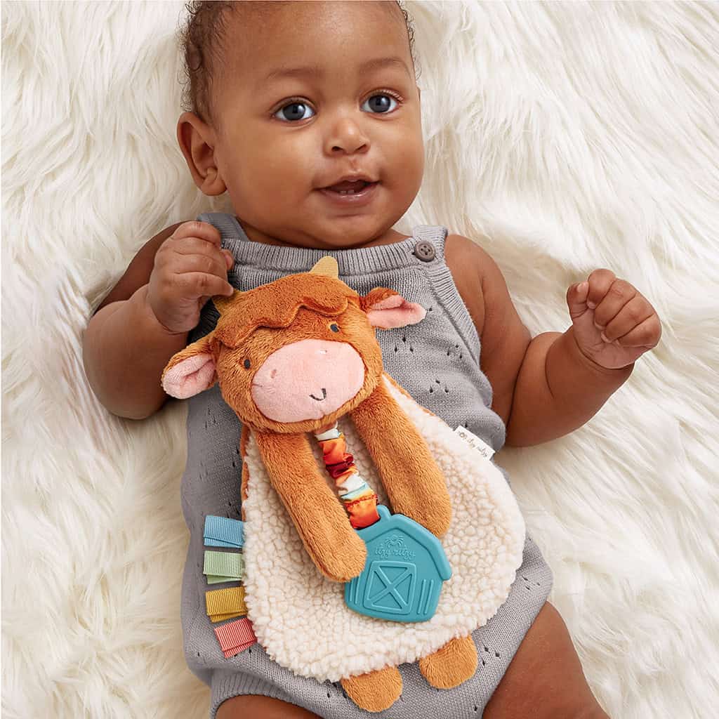 302801668_Itzy_Ritzy_Itzy_Lovey_Highland_Cow A baby in a gray romper lying on a white fluffy surface holds a plush toy shaped like a cow with a blue tag and colorful ribbons.