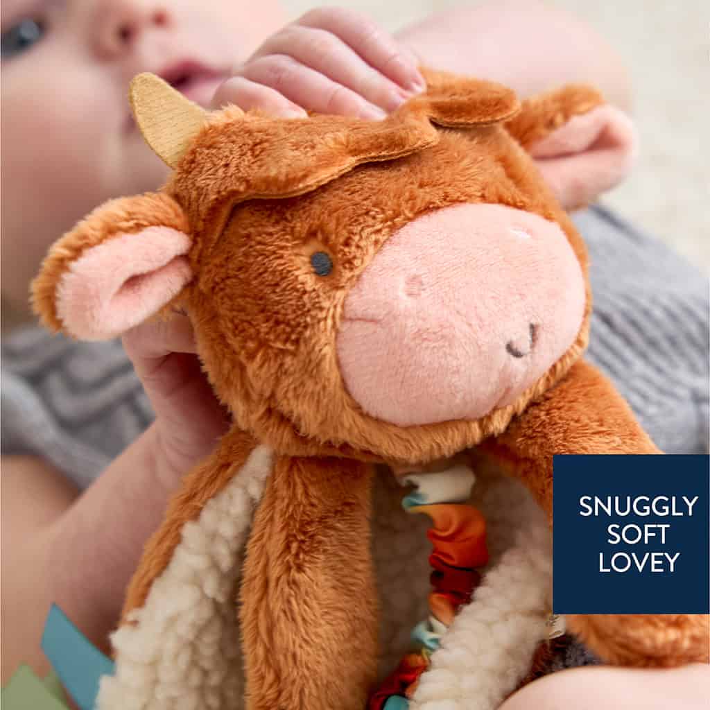 302801668_Itzy_Ritzy_Itzy_Lovey_Highland_Cow A baby holds a plush cow toy with a label reading "SNUGGLY SOFT LOVEY." The toy has brown fur, embroidered eyes, and a soft fabric underside with colorful ribbons.