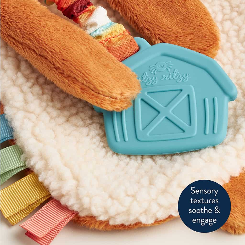 302801668_Itzy_Ritzy_Itzy_Lovey_Highland_Cow Close-up of a textured baby toy with a soft, fuzzy surface, colorful ribbon tags, and a blue silicone teether shaped like a barn. A label reads, "Sensory textures soothe & engage.