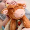 A close-up shows a baby holding a brown, plush toy cow. The toy has a round face, small horns, and floppy ears. The baby’s hand is visible, gripping the toy’s head.