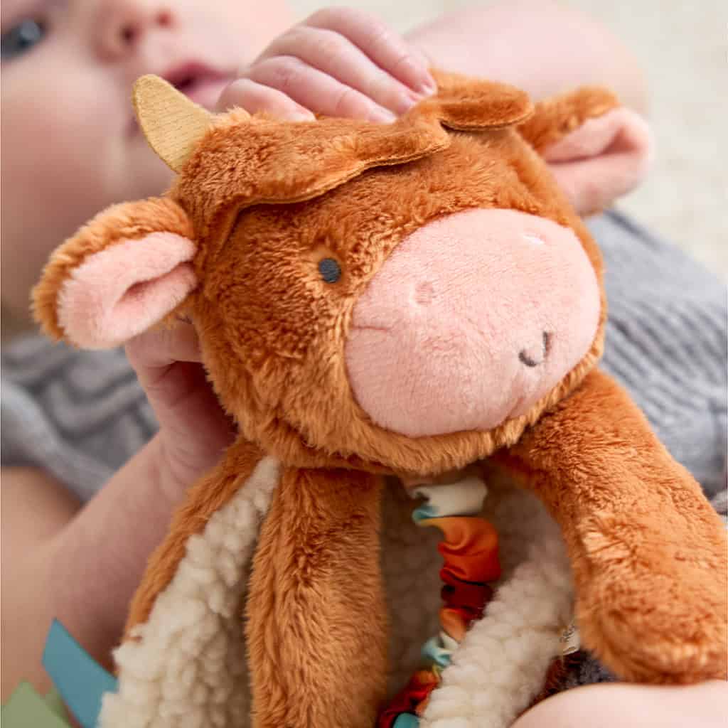 302801668_Itzy_Ritzy_Itzy_Lovey_Highland_Cow A baby holds a plush cow toy, which has a fluffy texture and small stitched eyes, ears, and a snout.