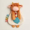 Plush toy of a smiling cow with brown fur, pink nose, and attached blue barn-shaped teether, adorned with colorful ribbon tabs on its left.
