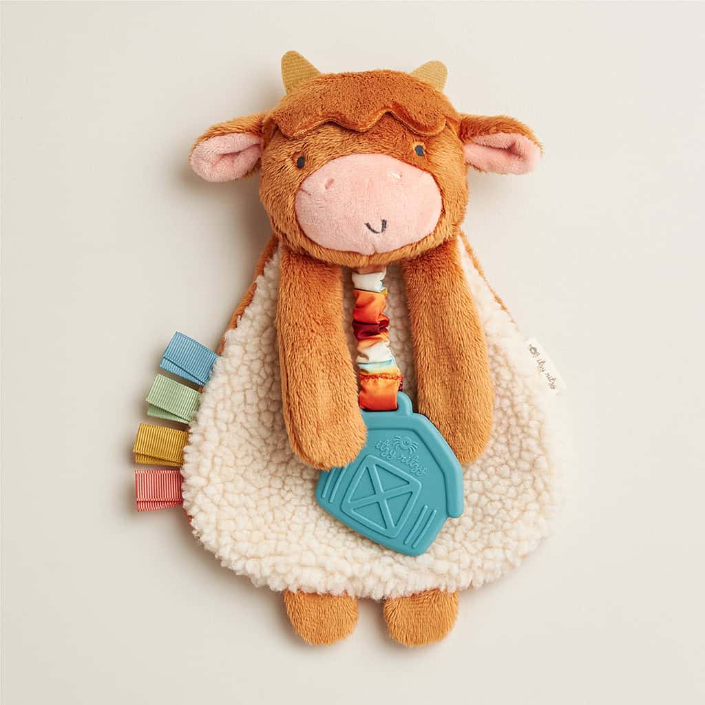 302801668_Itzy_Ritzy_Itzy_Lovey_Highland_Cow A plush toy shaped like a cow with a brown face and cream body, featuring colorful ribbon tags and an attached blue teething toy shaped like a barn.
