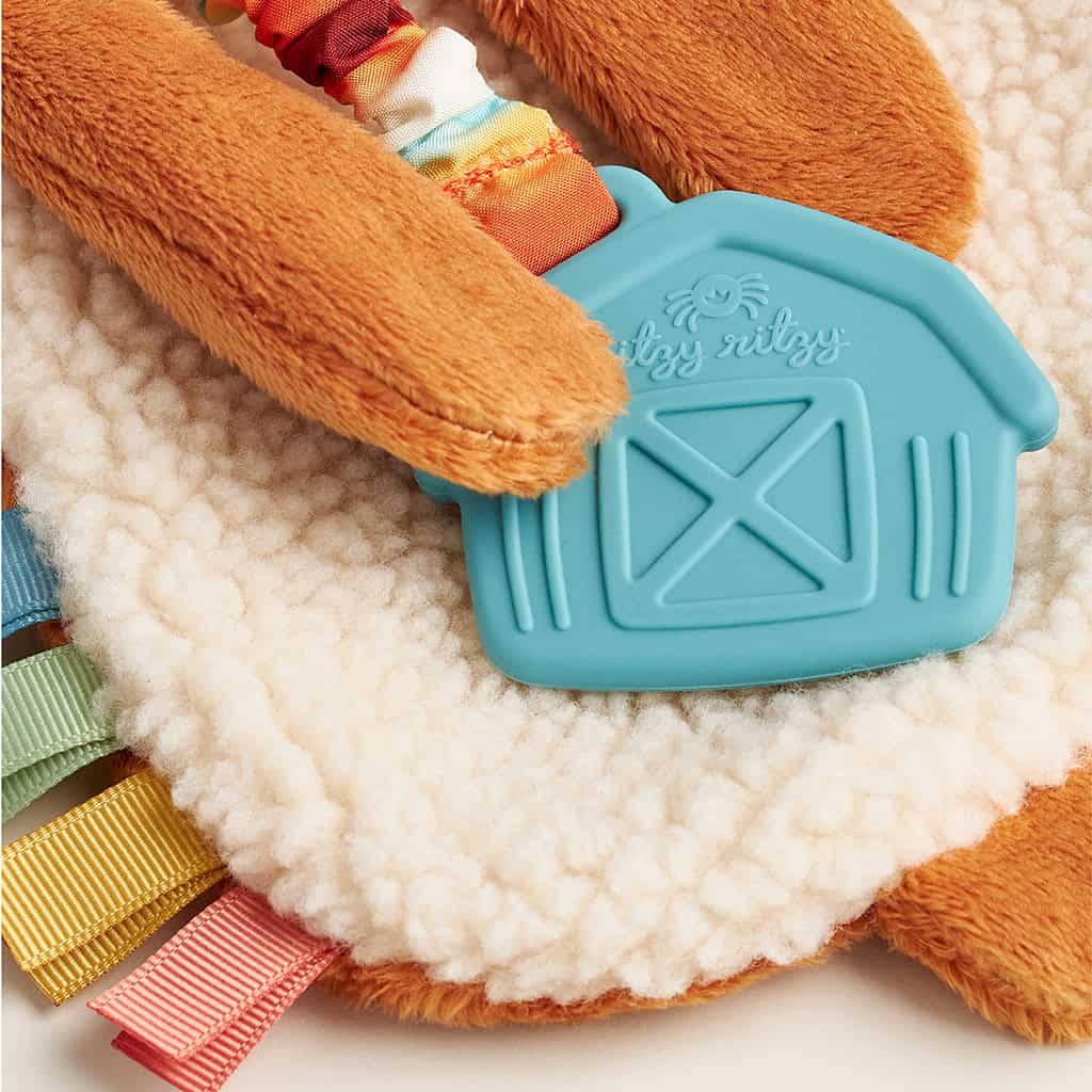 302801668_Itzy_Ritzy_Itzy_Lovey_Highland_Cow Close-up of a baby sensory toy with a blue silicone barn-shaped teether and colorful ribbons. The toy features soft, textured fabrics in various shades of brown and white.