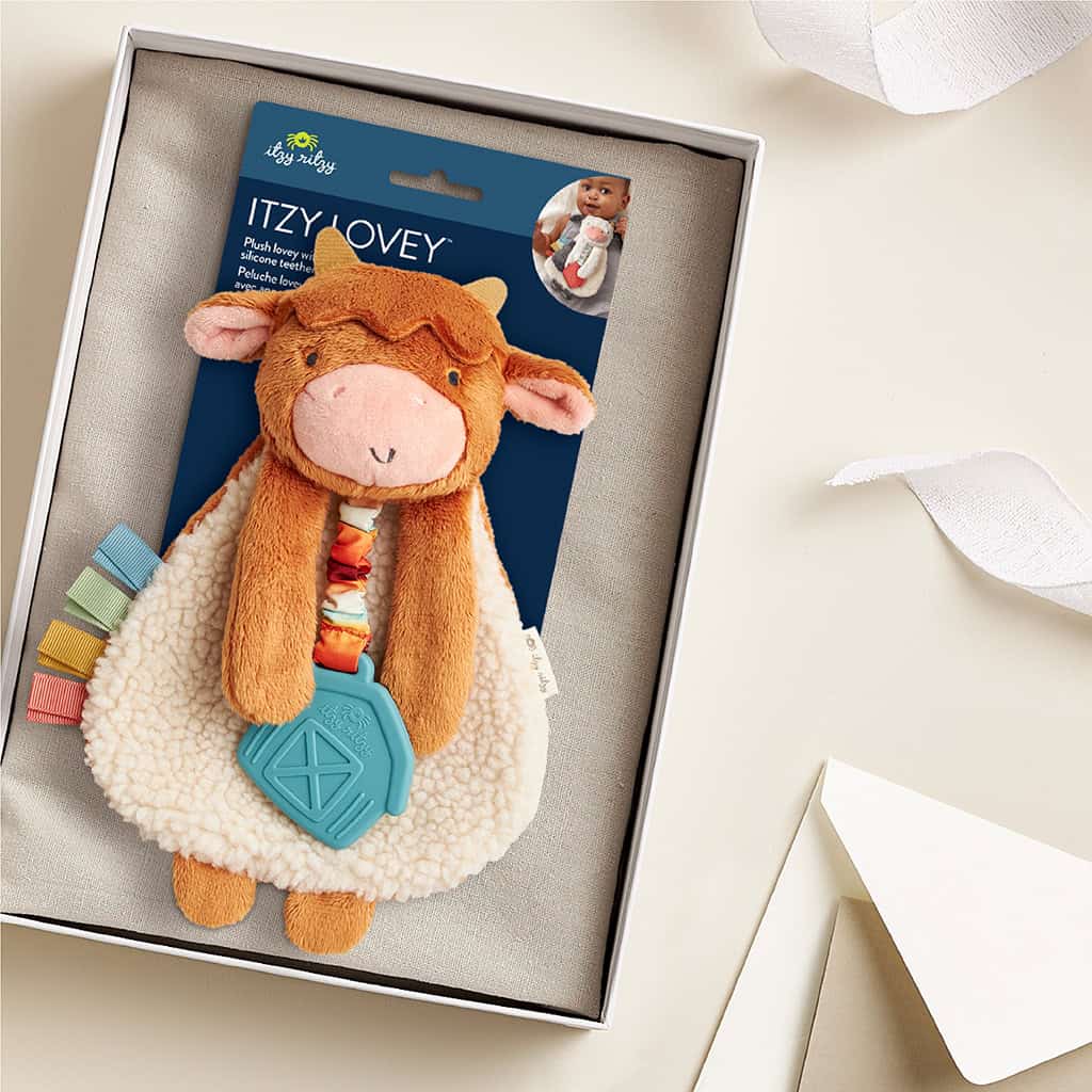 302801668_Itzy_Ritzy_Itzy_Lovey_Highland_Cow A plush toy packaged in a box labeled "Itzy Lovey" featuring a brown and white cow with a blue geometric teether and colorful ribbons attached. The background includes a white ribbon and an off-white envelope.