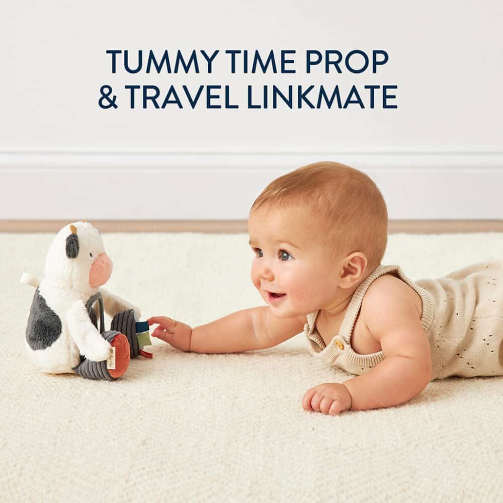 A baby lying on a beige carpet reaches toward a plush cow toy. Text above reads "Tummy Time Prop & Travel Linkmate.