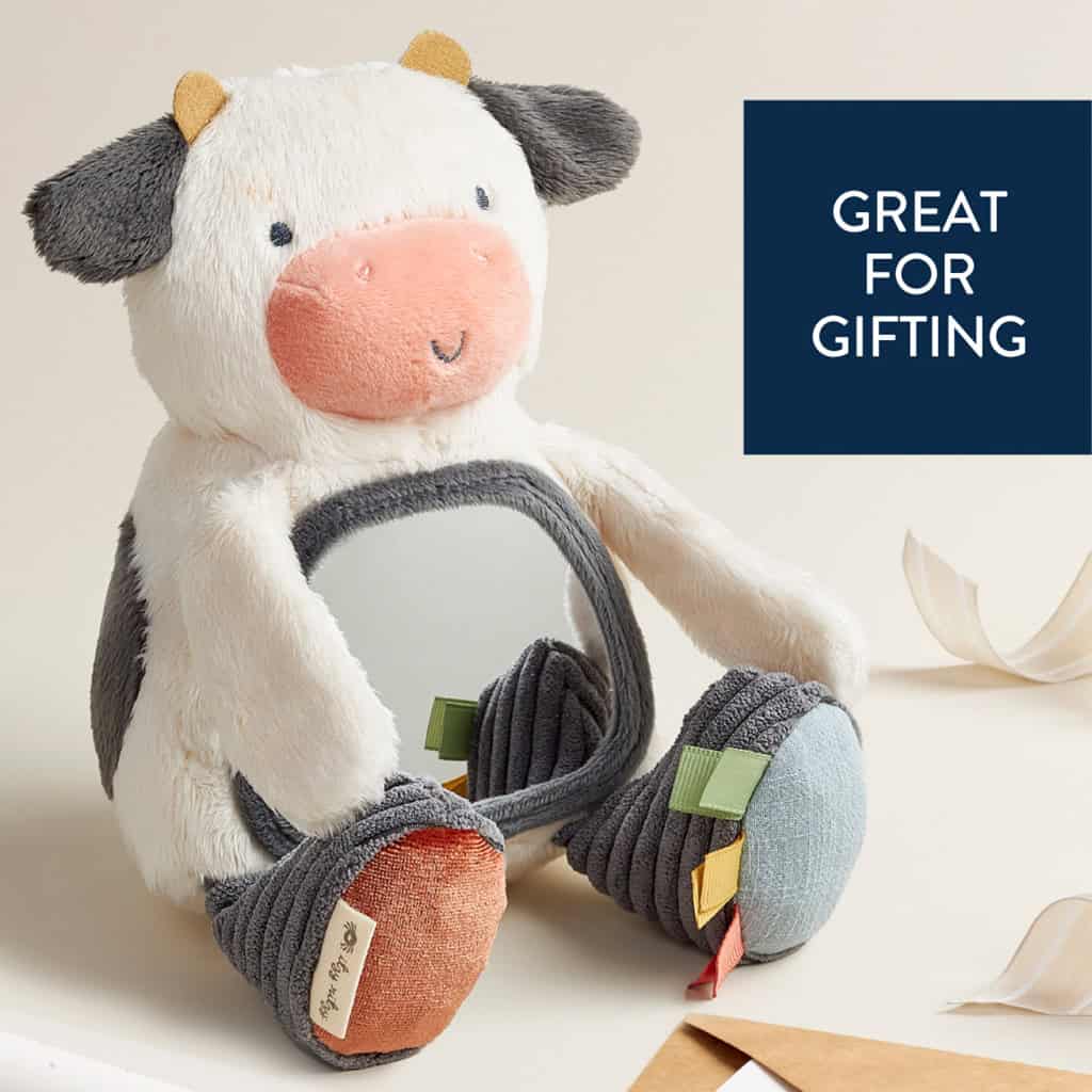 itzy-ritzy-306601562-itzy-bitzy-mirror-cow-2 A plush toy shaped like a cow with a mirror on its belly sits on a surface next to pieces of ribbon. A sign reads "Great for Gifting.