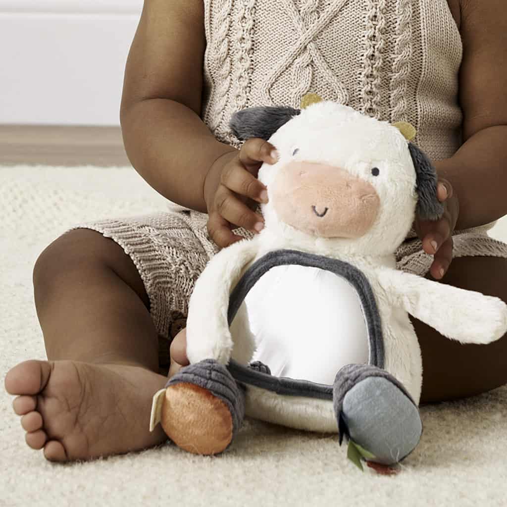 itzy-ritzy-306601562-itzy-bitzy-mirror-cow-4 A baby in a beige knitted outfit plays with a plush cow toy while sitting on a cream-colored rug.