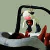 A baby lying in a gray car seat reaches up to touch a plush cow toy with a mirror and teething elements hanging above them.