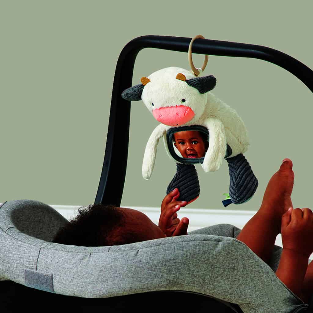 itzy-ritzy-306601562-itzy-bitzy-mirror-cow-5 A baby lying in a gray car seat reaches up to touch a plush cow toy with a mirror and teething elements hanging above them.