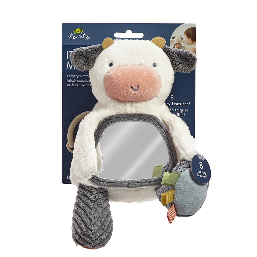itzy-ritzy-306601562-itzy-bitzy-mirror-cow-6 A plush cow toy with various textures and features, including a mirror on its tummy and a teether attached. Background packaging details sensory aspects.