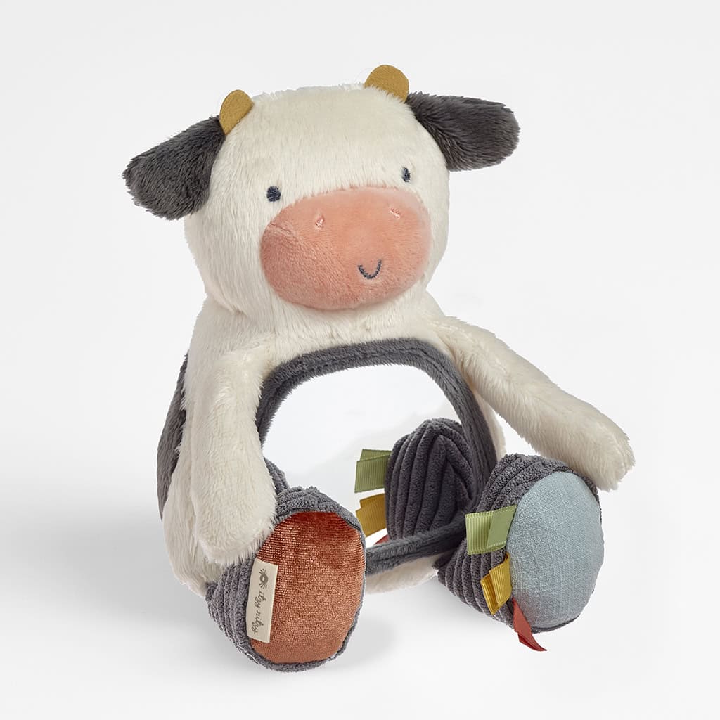 itzy-ritzy-306601562-itzy-bitzy-mirror-cow-7 Plush toy shaped like a cow with a round central opening, black and white fur, black ears, orange nose, and multicolored ribbons on the feet.