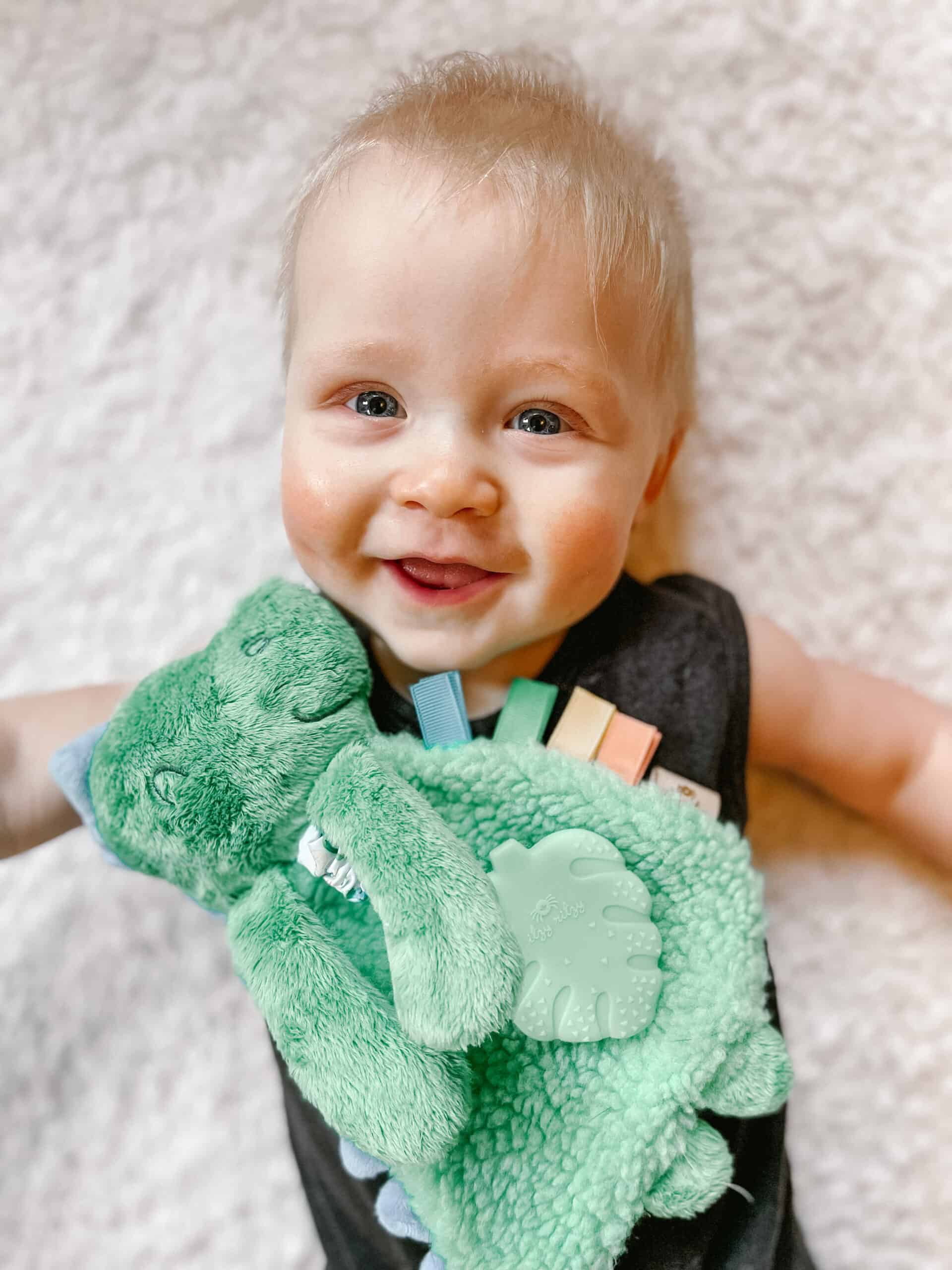 itzy-ritzy-lov8085-itzy-lovey-green-dino-10 A baby lies on a soft surface, smiling and holding a green stuffed toy with colorful tags.