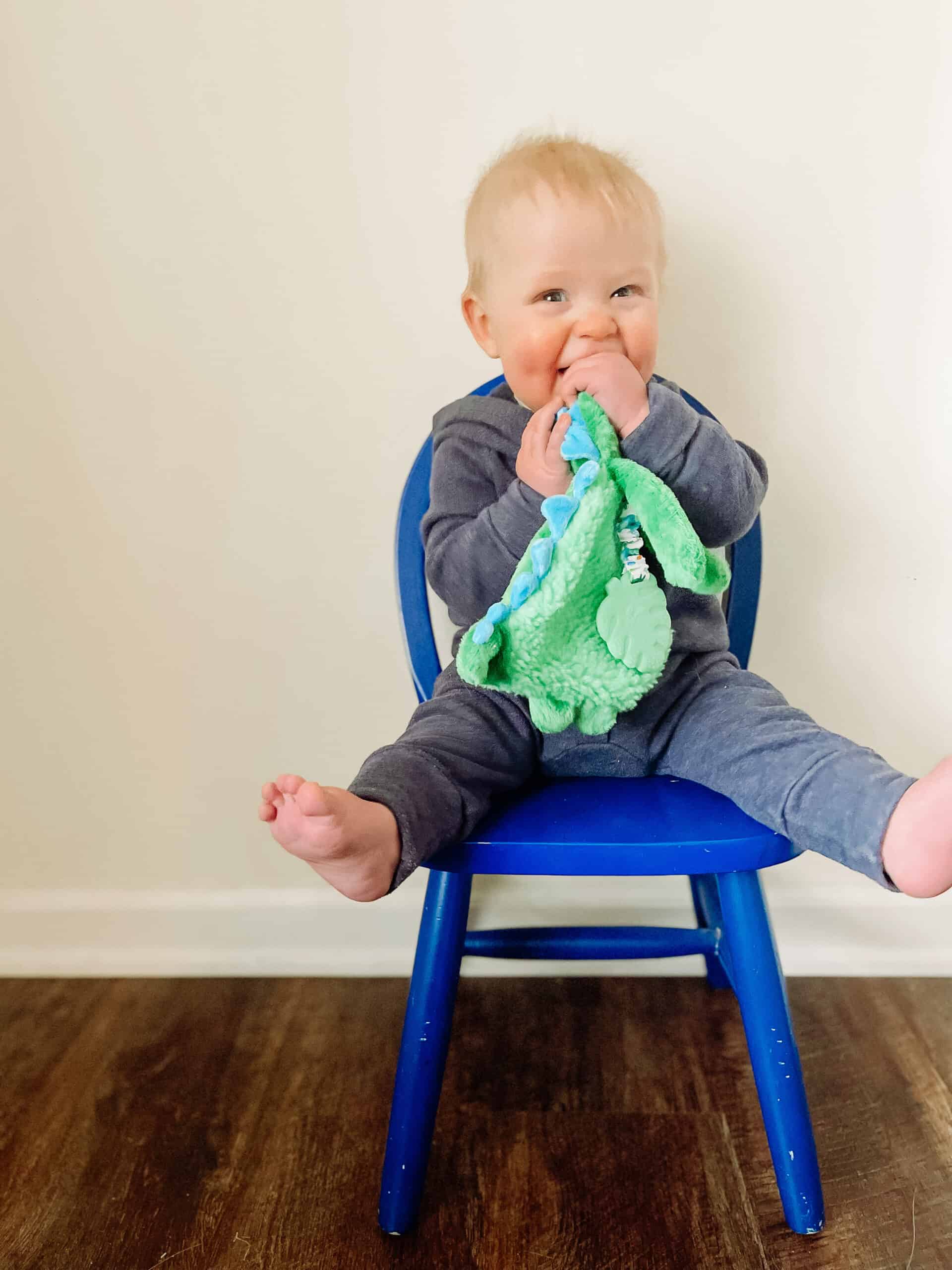 itzy-ritzy-lov8085-itzy-lovey-green-dino-11 A baby sits on a blue chair, wearing a grey outfit and chewing on a green blanket, with feet playfully off the edge of the chair.
