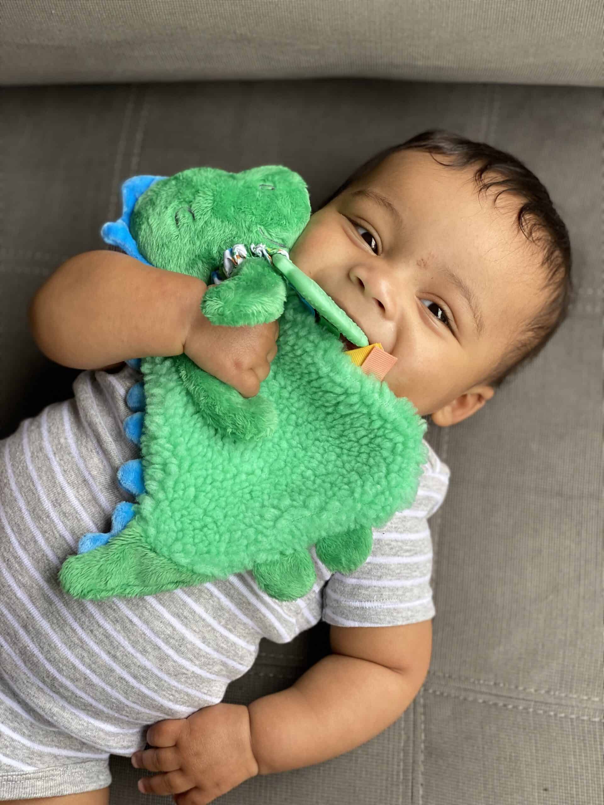 itzy-ritzy-lov8085-itzy-lovey-green-dino-12 A baby lies on a grey cushion, holding and chewing on a green plush dinosaur toy while wearing a grey and white striped onesie.