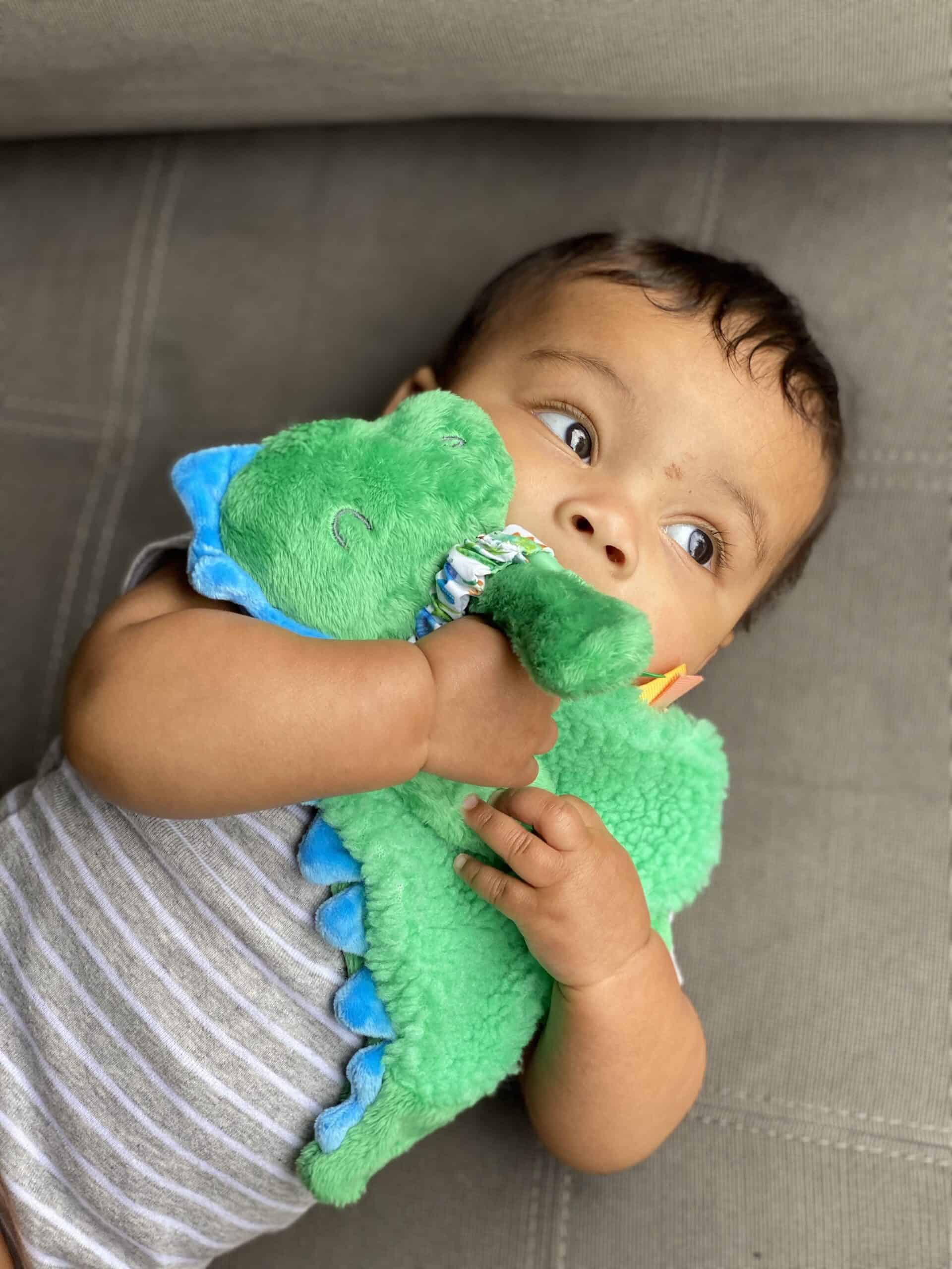 itzy-ritzy-lov8085-itzy-lovey-green-dino-13 A baby lies on a gray surface, wearing a gray and white striped onesie, holding a green dinosaur plush toy close to their face.