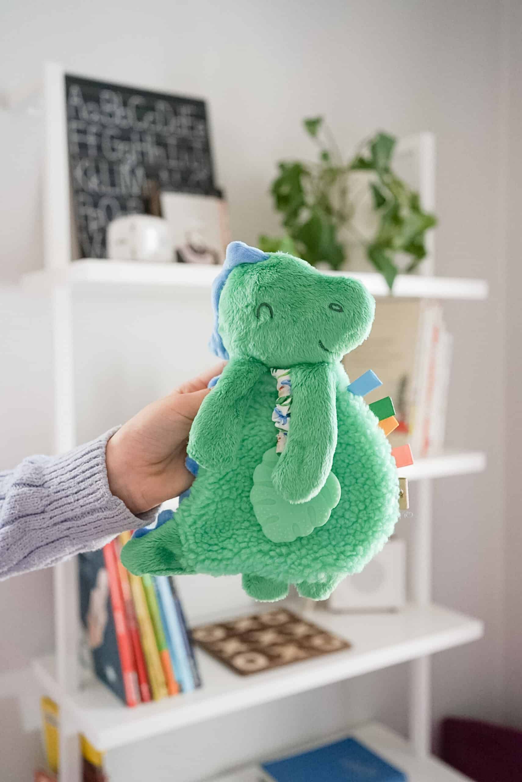 itzy-ritzy-lov8085-itzy-lovey-green-dino-5 A person holds a green dinosaur stuffed toy in a room with white shelves. The shelves contain books, plants, and decorative items.