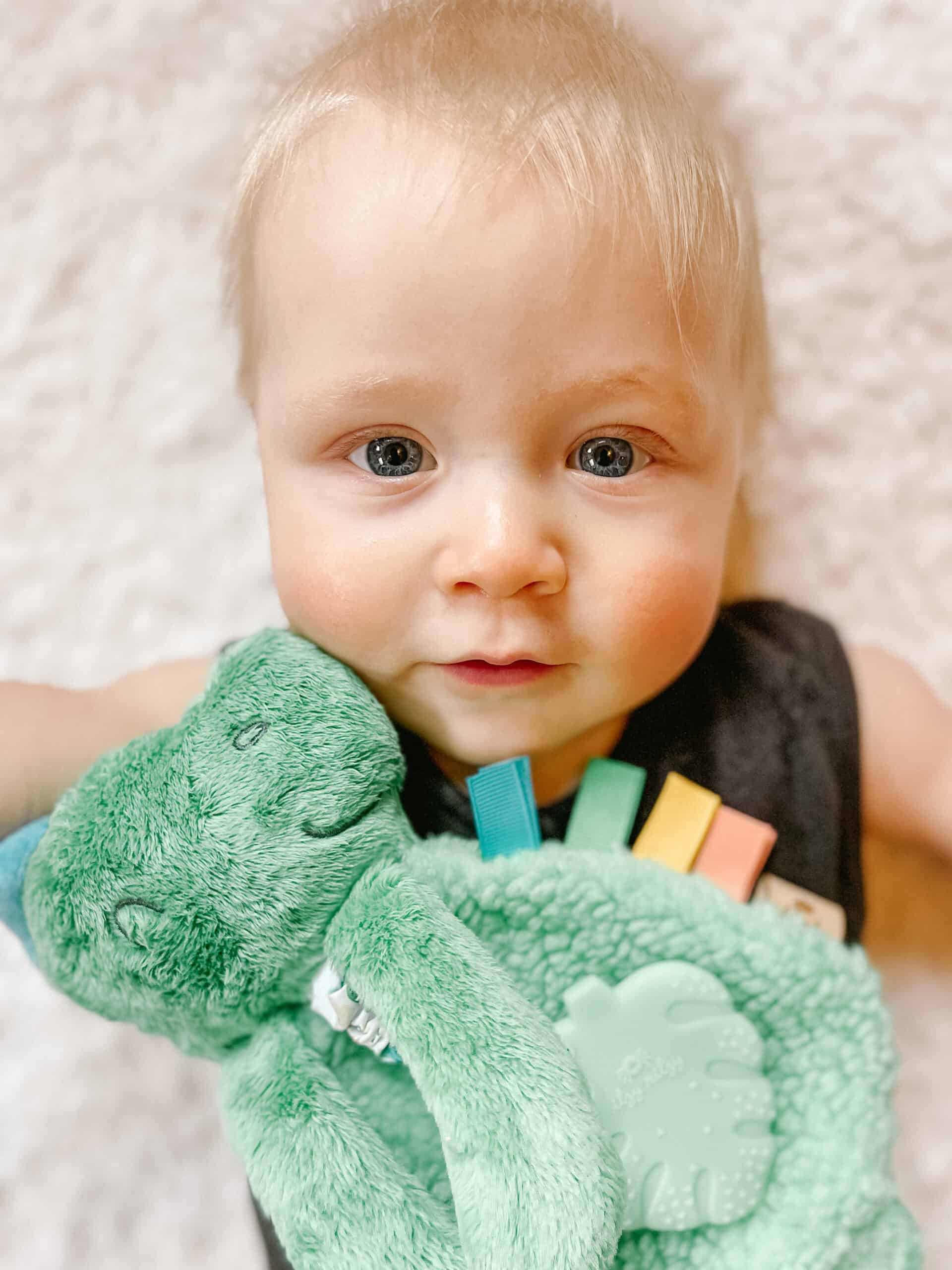 itzy-ritzy-lov8085-itzy-lovey-green-dino-6 A baby with light hair lies on a white blanket, holding a green plush toy. The baby is wearing a dark outfit and looking directly at the camera.