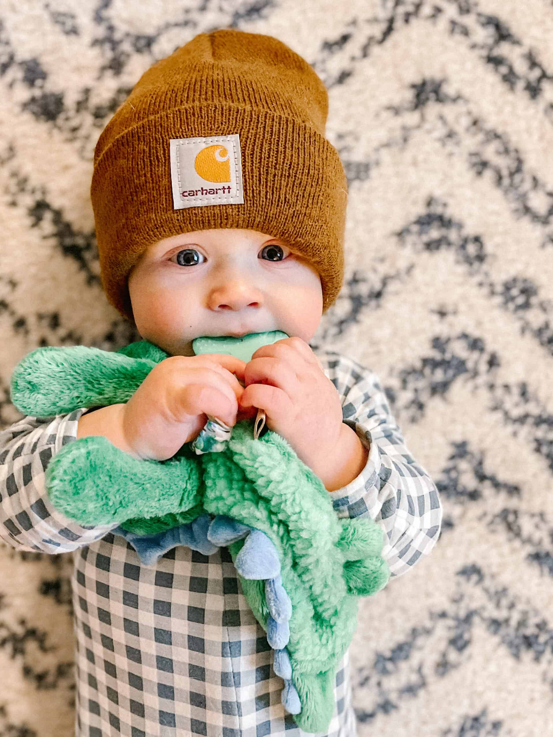 itzy-ritzy-lov8085-itzy-lovey-green-dino-7 A baby wearing a brown beanie and a checkered shirt holds a green plush toy and looks at the camera, standing in front of a patterned background.