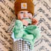 A baby, wearing a brown beanie and checkered outfit, smiles while holding and chewing on a green stuffed animal, standing in front of a patterned background.