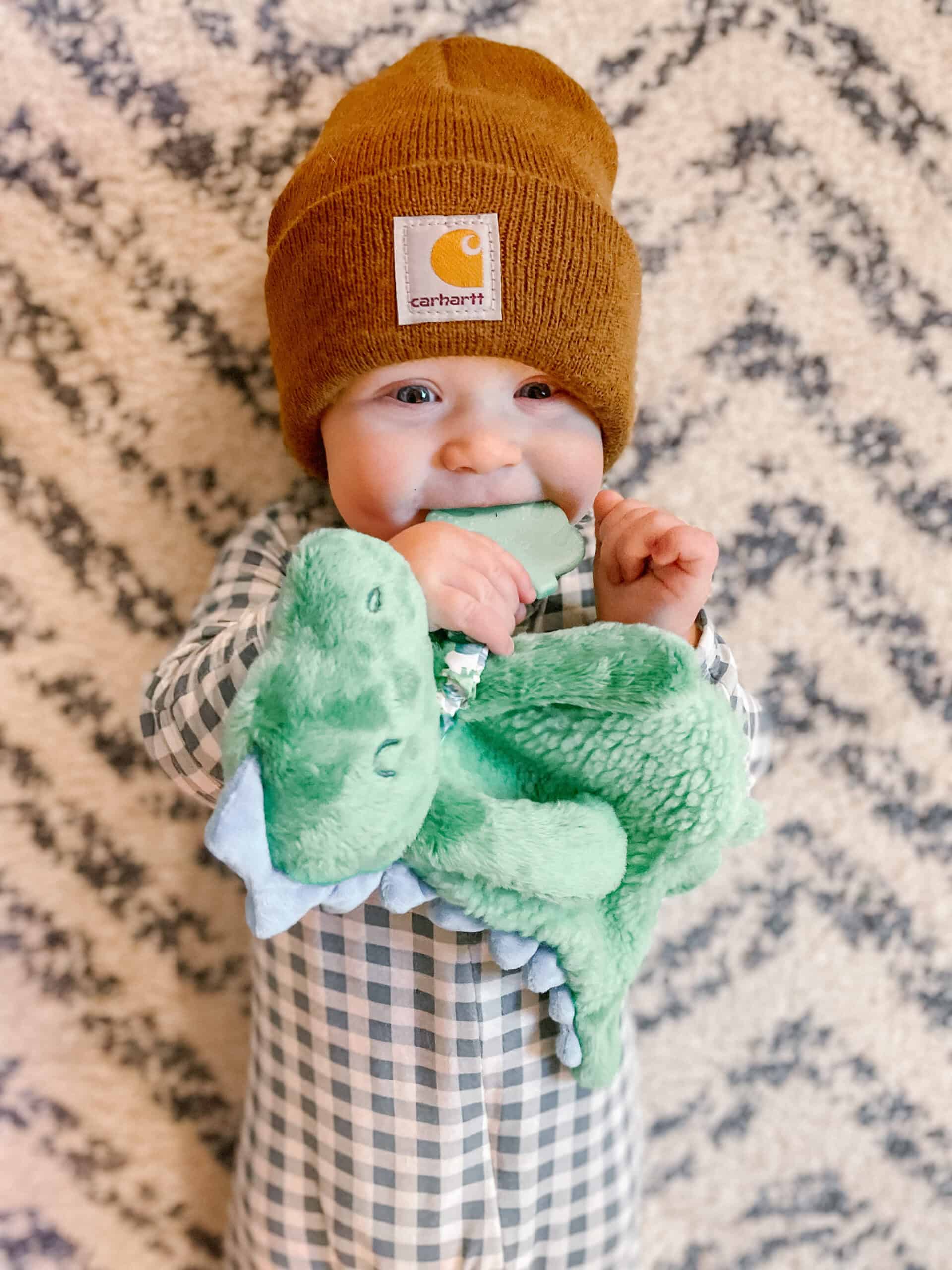 itzy-ritzy-lov8085-itzy-lovey-green-dino-8 A baby, wearing a brown beanie and checkered outfit, smiles while holding and chewing on a green stuffed animal, standing in front of a patterned background.