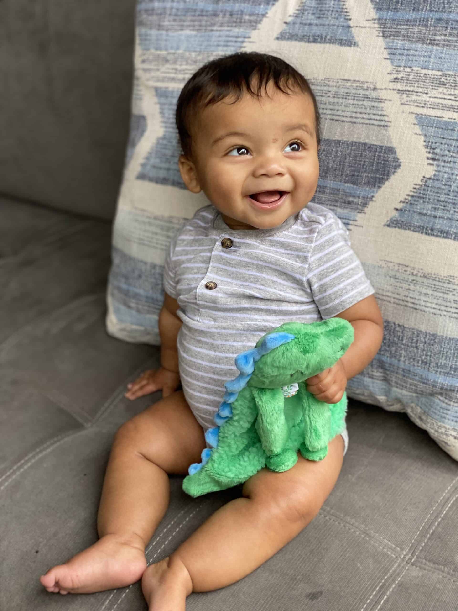 itzy-ritzy-lov8085-itzy-lovey-green-dino-9 A smiling baby sits on a couch holding a green dinosaur plush toy, with a patterned pillow behind them.