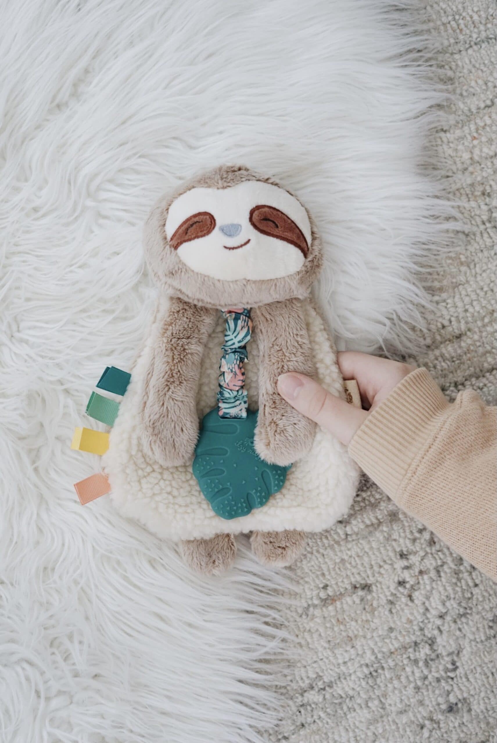 Processed with VSCO with a6 preset A hand holds a plush toy sloth with a white furry body and brown face, lying on a white fur rug. The toy features a teething ring and colorful fabric tags.