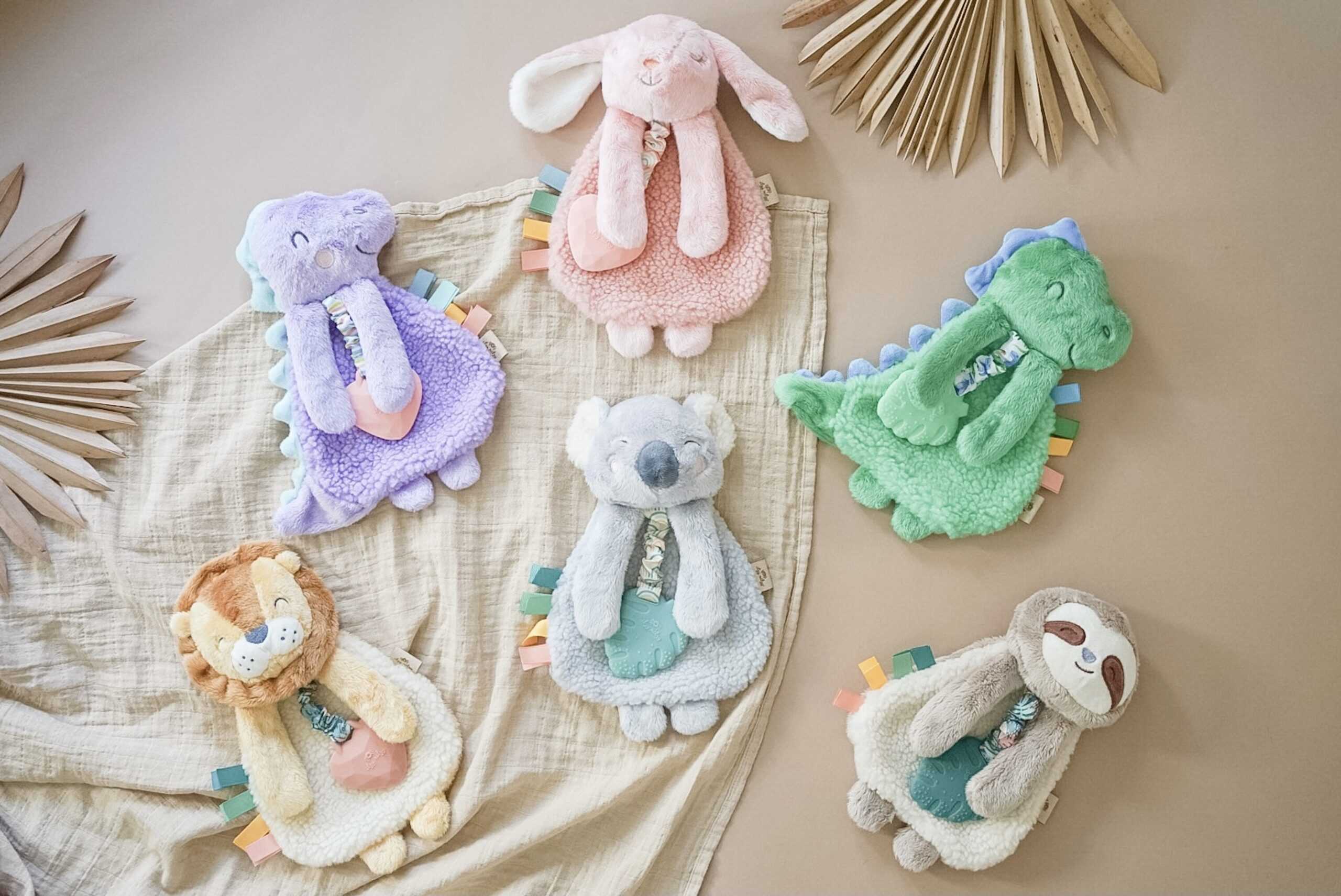 itzy-ritzy-lov8317-itzy-lovey-sloth-11 Six plush animal toys, including a lion, sloth, koala, and dinosaur, are arranged on a beige cloth with palm leaves in the background.