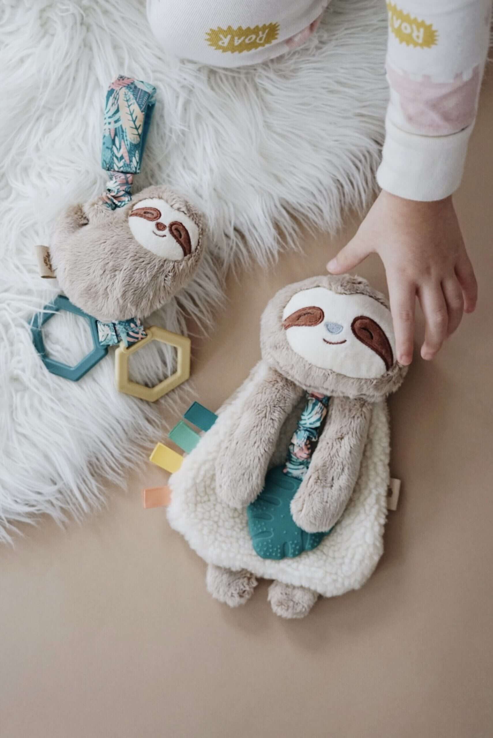 Processed with VSCO with a6 preset A child’s hand reaches for a plush sloth toy lying on a rug. The sloth toy has a smiling face and attached colorful ribbons. Another plush sloth toy with a plastic teether and geometric shapes lies nearby.